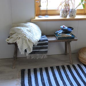 Può includere: Una panca di legno con una coperta bianca e un tappeto a strisce blu e bianche. La panca è decorata con un peluche e dei libri.
