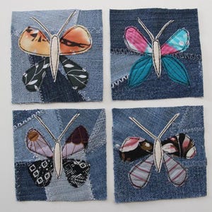 Op de afbeelding: Vier denim vierkanten met een patchwork-ontwerp, elk met een anders gekleurde vlinderapplicatie. De vlinders zijn gemaakt van verschillende stoffen en hebben een wit lichaam met een witte omtrek.
