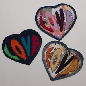 May include: Three denim heart-shaped patches with the word "LOVE" spelled out in different colors and patterns. The hearts are sewn with a denim border.