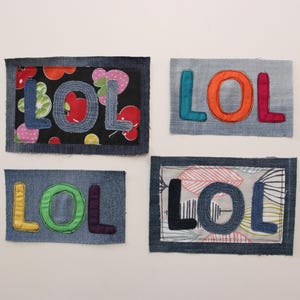 以下が含まれることがあります： 「LOL」と刺繍された、異なる色とパターンを持つデニムパッチが4つ。パッチはデニム生地で作られており、さまざまなカラフルな生地が縫い付けられています。
