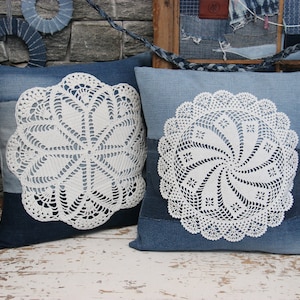 Op de afbeelding: Twee blauwe denim kussenhoezen met witte gehaakte doilies in het midden. De kussens liggen op een witgeschilderd houten oppervlak.
