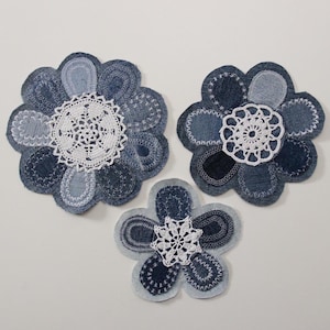 Può includere: Tre forme di fiori in denim blu con centri in pizzo bianco. I fiori sono realizzati in tessuto denim riutilizzato e sono cuciti insieme per creare un design unico e decorativo.