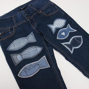 Op de afbeelding: Donkerblauwe spijkerbroek met verschillende lichtblauwe visapplicaties. De visontwerpen variëren, sommige met textuurpatronen en andere met een eenvoudige omtrek. De jeans hebben een klassiek ontwerp met vijf zakken en een knoopsluiting.