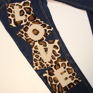 May include: Dark blue denim jeans with the word "LOVE" spelled out on the left leg. The letters are beige and are placed on top of leopard print patches. The jeans are laid flat on a white surface.
