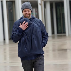 May include: A navy blue hooded jacket with a front-facing baby carrier insert, featuring ribbed cuffs, a full-length zip, and an adjustable drawstring. The jacket is paired with a grey knit beanie and dark grey trousers for a functional look.