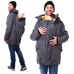 May include: A gray babywearing jacket with a hood and a front pocket. The jacket has a built-in pouch to carry a baby. The baby is wearing a yellow hat with a star design. The person is wearing a black beanie and blue jeans.