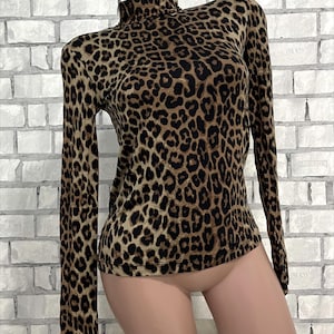 Puede incluir: Una camiseta de manga larga con cuello alto y estampado de leopardo. La camiseta presenta un cuello alto y una silueta ajustada. El estampado de leopardo consiste en manchas marrones y negras sobre un fondo beige. La camiseta se muestra en un maniquí.