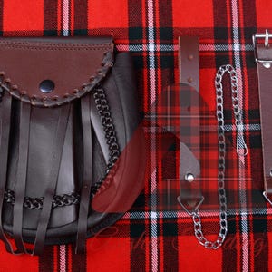 Pode incluir: Um sporran de couro marrom para kilt com uma corrente e uma alça. O sporran tem uma franja e é decorado com uma corrente. O sporran está sobre um fundo xadrez vermelho e preto.