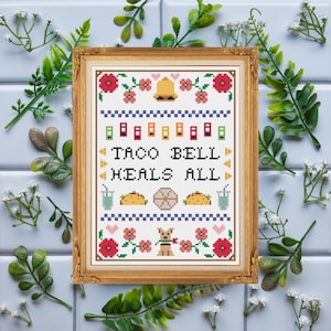 May include: A framed cross-stitch embroidery featuring the text "TACO BELL HEALS ALL" surrounded by pixelated motifs of a bell, flowers, hearts, tacos, drinks, and a dog, all rendered in a colorful palette within a gold-toned decorative frame.