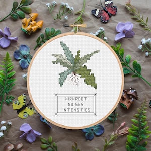 May include: A cross-stitch embroidery hoop featuring a green and light green fern design with visible roots. The text "NIRNROOT NOISES INTENSIFIES" is below the fern. The background includes decorative flowers, butterflies, and mushrooms.