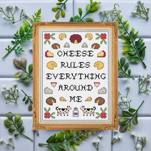 May include: A framed cross-stitch piece with the text "CHEESE RULES EVERYTHING AROUND ME." The design includes various cheese illustrations, cows, and floral accents. The frame is gold-colored, and the background is white.