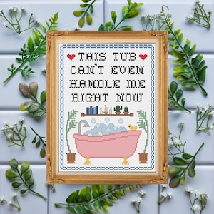 May include: Cross-stitch artwork in a gold frame. The design shows a pink bathtub with bubbles and a rubber duck, with the text "THIS TUB CAN'T EVEN HANDLE ME RIGHT NOW". The artwork is surrounded by green foliage and white flowers.