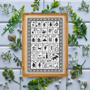 Puede incluir: Un muestrario bordado enmarcado con un marco dorado. El diseño en blanco y negro presenta un alfabeto con imágenes y números con temática de Halloween. El muestrario está rodeado de hojas verdes y flores blancas.