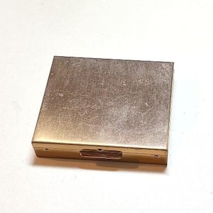 May include: A square, gold-toned metal compact case with a hinged lid and a latch closure. The surface of the lid features a brushed, textured finish with visible fine scratches and signs of wear across the metallic top panel.