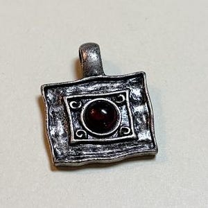 May include: A rectangular silver-toned metal pendant featuring a textured, uneven surface and a central square frame. The frame contains a smooth, dark red circular cabochon stone and small decorative scrollwork details at each corner.