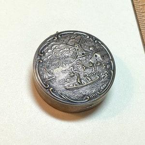 May include: Silver-toned round trinket box with an embossed design of a windmill, houses, and a boat with people on a river. The box has a dark gray background and a silver-toned rim.
