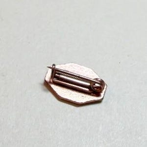 May include: A small, copper-colored pin with a geometric shape. The pin has a safety clasp on the back and a small, decorative front. The pin is likely made of metal and is suitable for attaching to clothing or accessories.