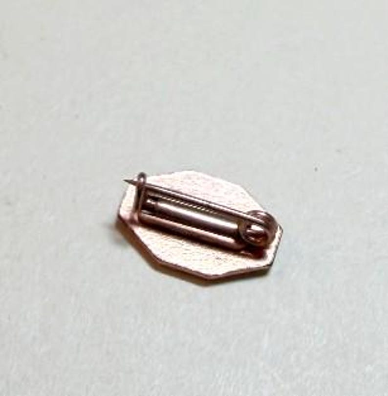 May include: A small, copper-colored pin with a geometric shape. The pin has a safety clasp on the back and a small, decorative front. The pin is likely made of metal and is suitable for attaching to clothing or accessories.