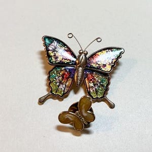 May include: A butterfly-shaped brooch with metallic edges and wings filled with multicolored glitter in shades of green, purple, and red. Below it sits a smaller, separate butterfly-shaped pin back featuring a solid, light tan marbled finish.
