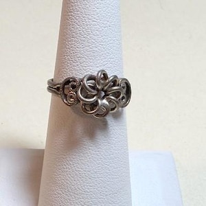 May include: A silver-toned ring with an intricate floral design. The ring features a central flower-like cluster and decorative openwork on the band. The ring is displayed on a white, cone-shaped stand.