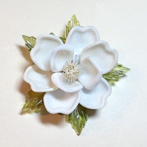 May include: A white flower brooch with layered petals and a textured center. The brooch is accented with translucent green leaves. This floral accessory is suitable for adding a touch of nature-inspired elegance to any outfit.