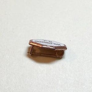 May include: A small, vintage pin with a white enamel surface and the text "WILDWOOD N.J." in black. The pin has a copper-colored metal frame and a safety pin closure. The pin is likely a souvenir or collectible item.