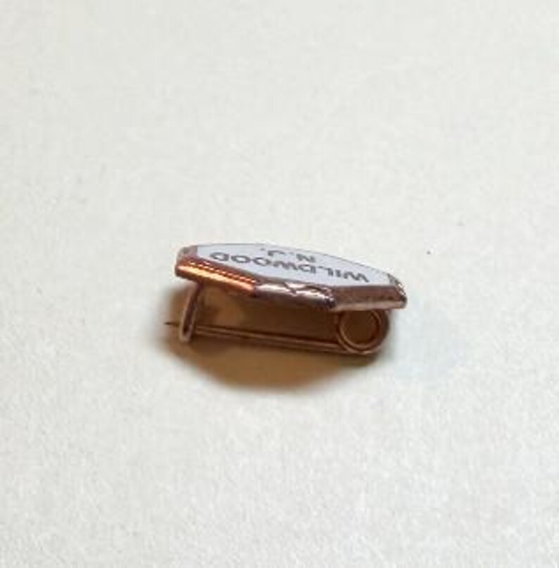 May include: A small, vintage pin with a white enamel surface and the text "WILDWOOD N.J." in black. The pin has a copper-colored metal frame and a safety pin closure. The pin is likely a souvenir or collectible item.