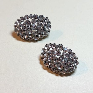 May include: Two oval-shaped, silver-toned earrings encrusted with numerous clear, sparkling rhinestones. The rhinestones are of varying sizes, creating a textured surface. The earrings are displayed on a light-colored background.