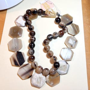 May include: A statement necklace featuring a mix of geometric and round beads in shades of brown, cream, and black. The necklace includes large, hexagonal beads and smaller, spherical beads, creating a bold and textured design. The tag reads "Sunshine Denda".