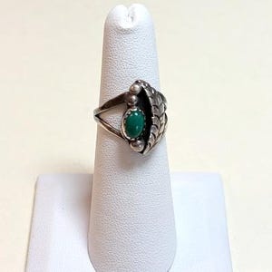 May include: A silver ring with a leaf design and a central oval-shaped green stone. The ring features a detailed leaf on one side and two small spheres on either side of the green stone. The ring is displayed on a white finger form.