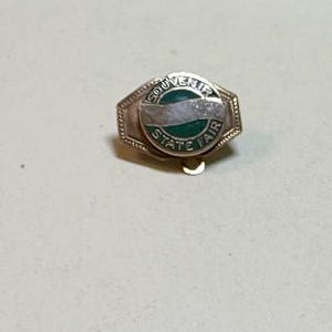 May include: A circular metal pin with a green and silver enamel finish. The center features a horizontal silver band across a green background, with the text SOUVENIR STATE FAIR embossed in silver lettering along the circular border of the pin.