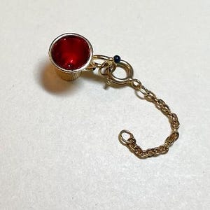 May include: A gold-colored metal wine bottle stopper with a bright red, round top. A small gold chain is attached to the stopper, ending in a clasp. A small blue bead is on the chain.