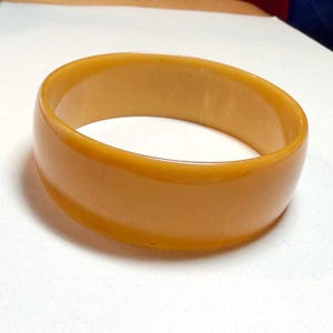 May include: A solid, smooth, mustard-yellow bangle bracelet with a wide, rounded profile. The accessory features a glossy, uniform finish throughout its entire surface, showcasing a simple, continuous circular band design with no additional embellishments.