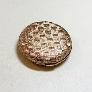 May include: A gold-toned, round compact with a textured, woven pattern. The compact has a hinged lid and a small clasp. The overall design is vintage and elegant, suggesting a cosmetic or jewelry case.