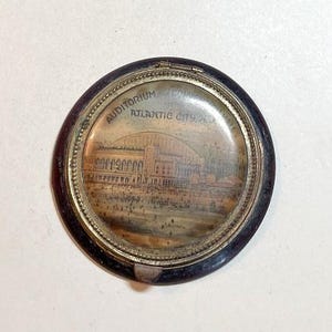 May include: A round, wooden, black compact with a gold rim and a glass insert featuring a black and white image of the Auditorium in Atlantic City, New Jersey.