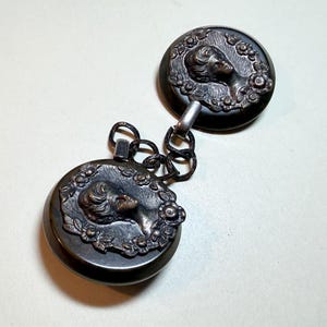 May include: Antique silver-toned clasp with two circular components connected by a chain. Each component features a profile of a woman surrounded by a floral wreath. The clasp is likely for a necklace or garment.