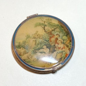 May include: A round, antique compact with a detailed painted scene under a clear cover. The image depicts figures in a pastoral setting with a waterfall and trees. The compact has a gold-colored metal frame with a blue enamel border.