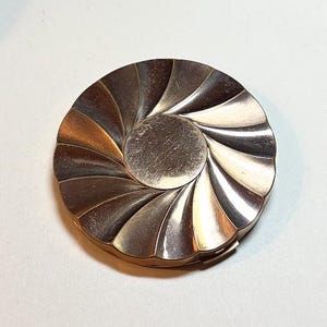 May include: A vintage silver-toned compact mirror with a spiral design. The compact has a circular shape with a central disc and radiating, curved segments. The metal has a brushed finish, and the compact appears to be hinged.