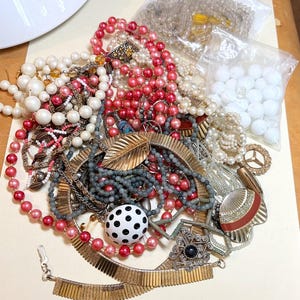 May include: A collection of vintage necklaces and jewelry in various styles and colors. The assortment includes beaded necklaces in shades of red, white, and gray, along with gold-toned metal pieces and a black and white polka dot bead.