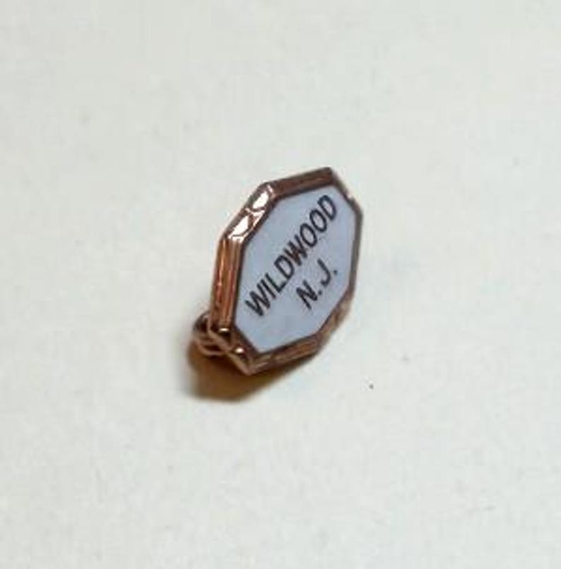 May include: A small, octagonal pin with a white center and a gold-colored border. The pin features the text "WILDWOOD N.J." in black, with a small loop for attachment. The pin is likely a souvenir or collectible item.