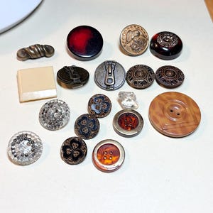 May include: A collection of assorted buttons in various shapes, sizes, and materials. The set includes round, square, and oval buttons featuring metallic, clear, amber, and black finishes, some with intricate embossed patterns and zipper motifs.