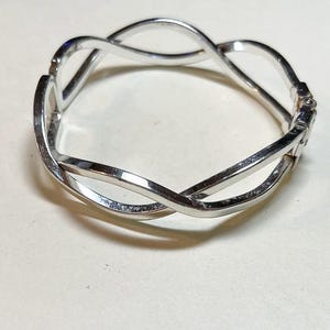 May include: A silver-colored bangle bracelet with an interwoven design. The bracelet features a clasp closure and a polished finish. The design is a continuous loop, creating an elegant and modern look. The bracelet is approximately 2.5 inches in diameter.