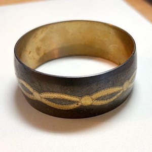 May include: A wide, antique-style bracelet with a dark exterior and a gold-colored interior. The bracelet features a repeating gold-colored chain design around the exterior. The bracelet is made of metal and has a vintage aesthetic.