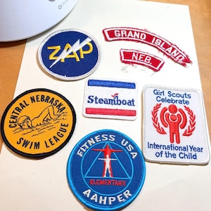 May include: A collection of seven embroidered fabric patches featuring various designs: a blue ZAP patch, red Grand Island and NEB patches, a yellow Central Nebraska Swim League patch, a Steamboat patch, a Fitness USA AAHPER patch, and a Girl Scouts Celebrate patch.