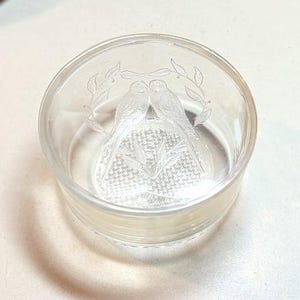 May include: A clear glass dish with a detailed etched design. Two birds are depicted facing each other, surrounded by leaves. The base of the dish has a textured pattern. The dish is round and has a slightly raised rim.