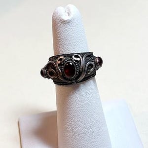 May include: A silver ring with an ornate design featuring dark red gemstones. The ring has a textured band with swirling patterns and is displayed on a white finger form. The gemstones are oval-shaped and set within the design.