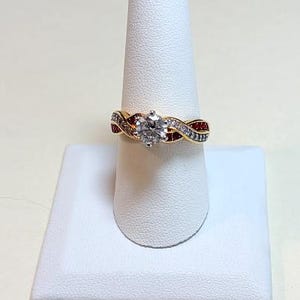 May include: A gold-toned ring with a large, clear, heart-shaped center stone. The band features a twisted design with alternating rows of small white and red stones. The ring is displayed on a white ring holder.