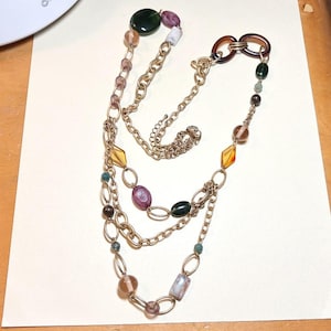 May include: A multi-strand necklace featuring gold-tone metal chains interspersed with various beads in shades of green, purple, amber, and brown. The design includes oval and rectangular beads, along with circular links, creating a layered appearance.