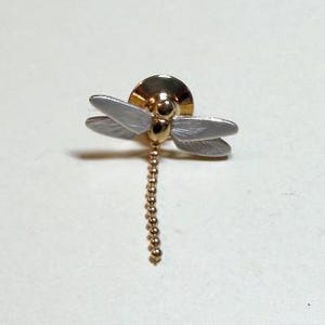 May include: A dragonfly lapel pin featuring a polished gold-tone body and head with four matte white wings. A long, thin gold-tone ball chain extends downward from the body, creating a dangling effect for this decorative accessory piece.
