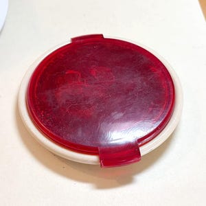 May include: A round, red plastic object with a white rim. The red center has a slightly textured appearance. Two small, red tabs extend from the red center. The object is circular and appears to be a lid or cover.